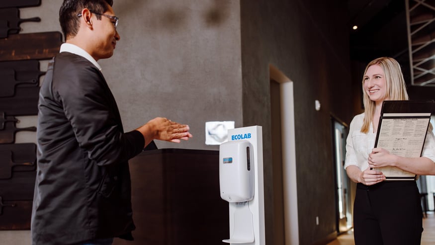 Restaurant hostess greeting a customer who is sanitizing their hands using Ecolab hand sanitizer from a Nexa Hand Hygiene Dispenser.