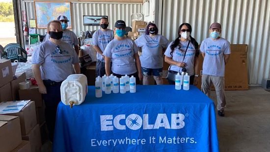 A group of mask-wearing volunteers wearing "Ecolab Volunteers" t-shirts gathered around a table, with a tablecloth that reads "Ecolab Everywhere It Matters," and disinfectant bottles atop the table